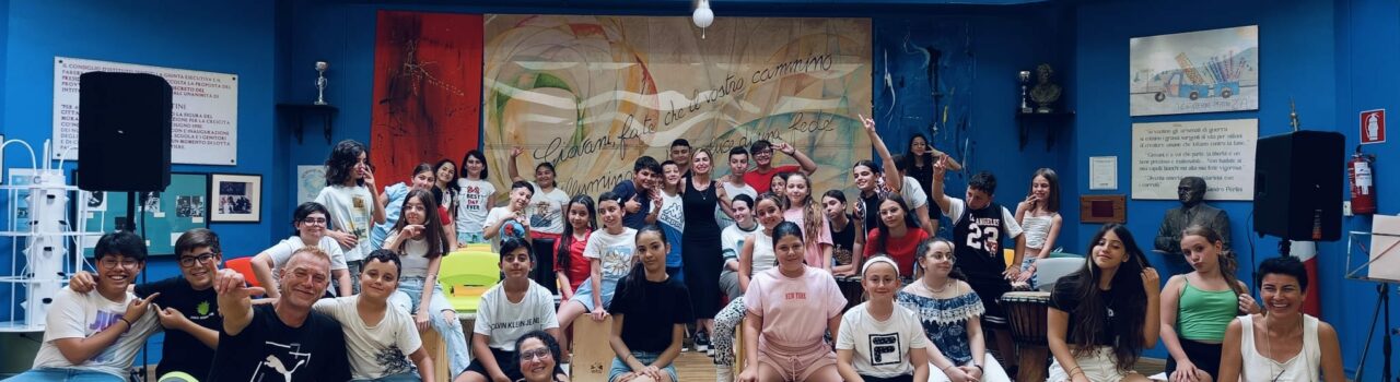 alunni e formatori in aula magna della Pertini dopo l'esibizione.L'immagine associata al corso presenta un collage di tamburi tradizionali con decorazioni artistiche. Il titolo "La voce del tamburo" è scritto in grandi caratteri neri su sfondo bianco. Sopra il titolo, sono riportati i loghi dei patrocini e la data di inizio del corso. Al centro, il logo dell'ICS Sperone Pertini.