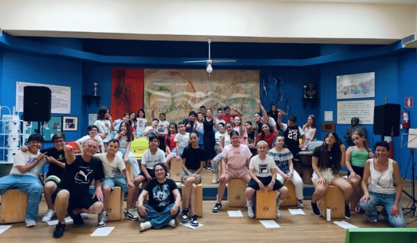 alunni e formatori in aula magna della Pertini dopo l'esibizione.L'immagine associata al corso presenta un collage di tamburi tradizionali con decorazioni artistiche. Il titolo "La voce del tamburo" è scritto in grandi caratteri neri su sfondo bianco. Sopra il titolo, sono riportati i loghi dei patrocini e la data di inizio del corso. Al centro, il logo dell'ICS Sperone Pertini.